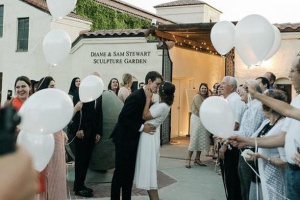 Photo of wedding couple kissing in the center of a group of guests who are holding balloons and smiling