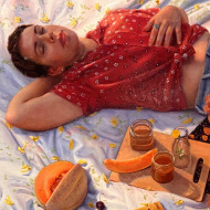 the upper half of a woman laying on a blanket in a red shirt, with a cantaloupe and slices of it laying on her right