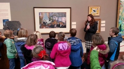 Group of elementary school children gathered in front of a painting on a gray wall, with a docent talking to them
